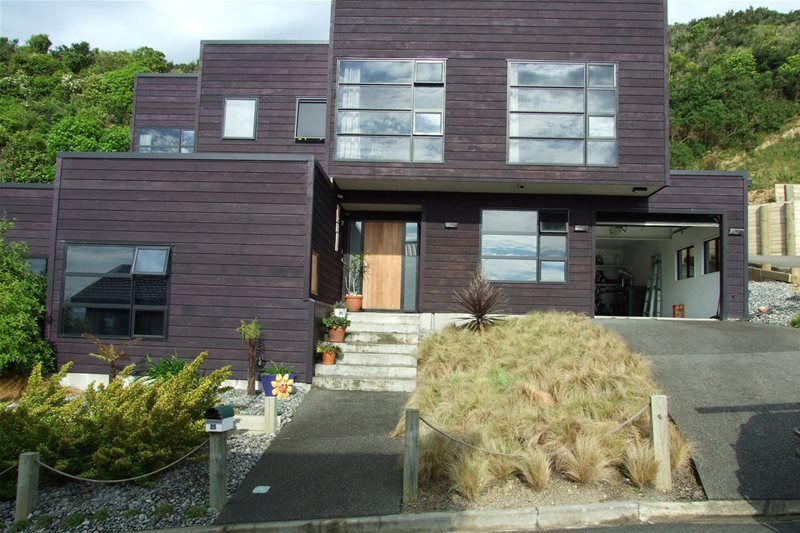 Modern dark wood house on a hillside with stairs, large windows, and dry ornamental grass in front.