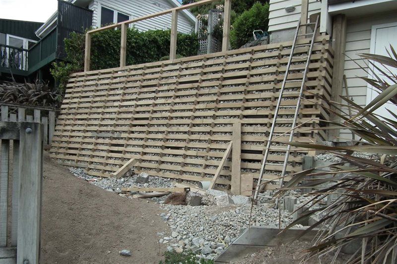 Retaining wall made of stacked wooden beams beside a sloped gravel path and house exterior