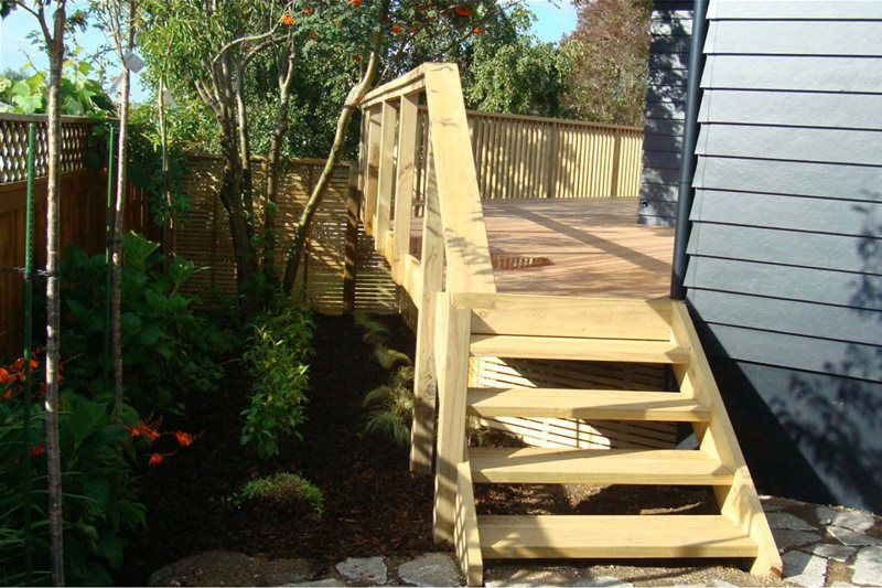 Wooden deck stairs beside a gray house, leading to a fenced backyard garden.