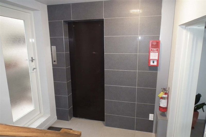 Small elevator lobby with gray tiled wall, dark elevator doors, wall mirror, and fire extinguisher.