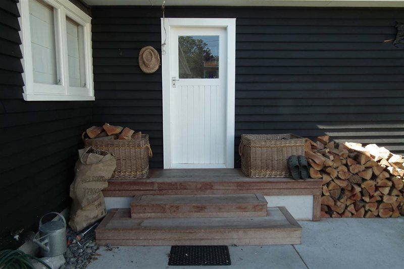 Front porch with white door, steps, firewood stacks, and a watering can beside a dark house wall