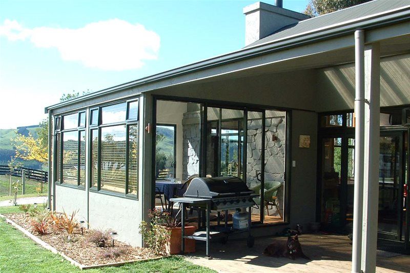 Glass sunroom and patio beside a house, with a grill and chairs overlooking a garden.