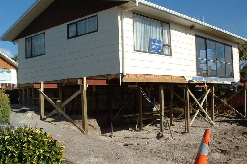 Raised house on wooden stilts during construction, with gravel ground and an orange traffic cone nearby