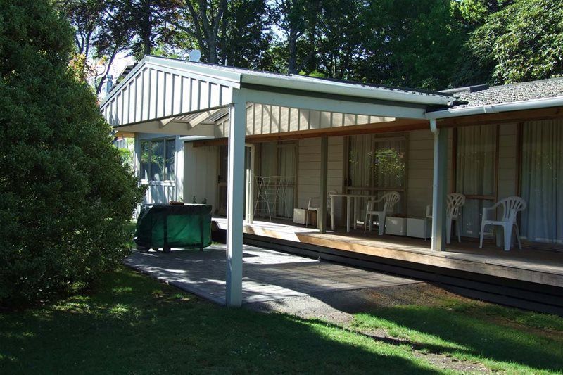 Small house porch with white posts, green bins, and outdoor chairs beside a lawn and trees.
