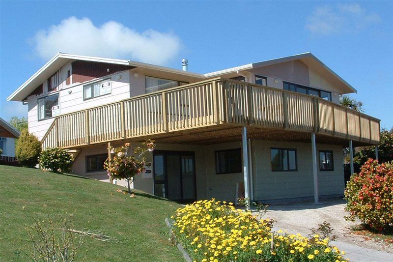 Modern two-story house with a wooden deck, grassy lawn, and yellow flowers under a blue sky