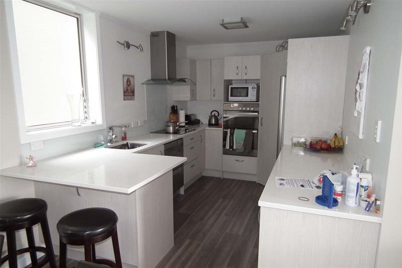 Modern white kitchen with island, dark flooring, built-in ovens, and a bright window over the sink.