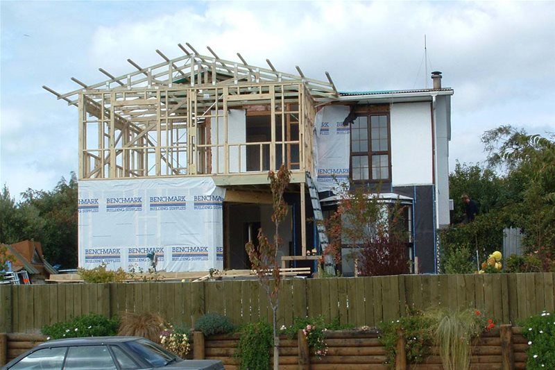 Partially built modern house with exposed wooden framing and scaffolding behind a fence