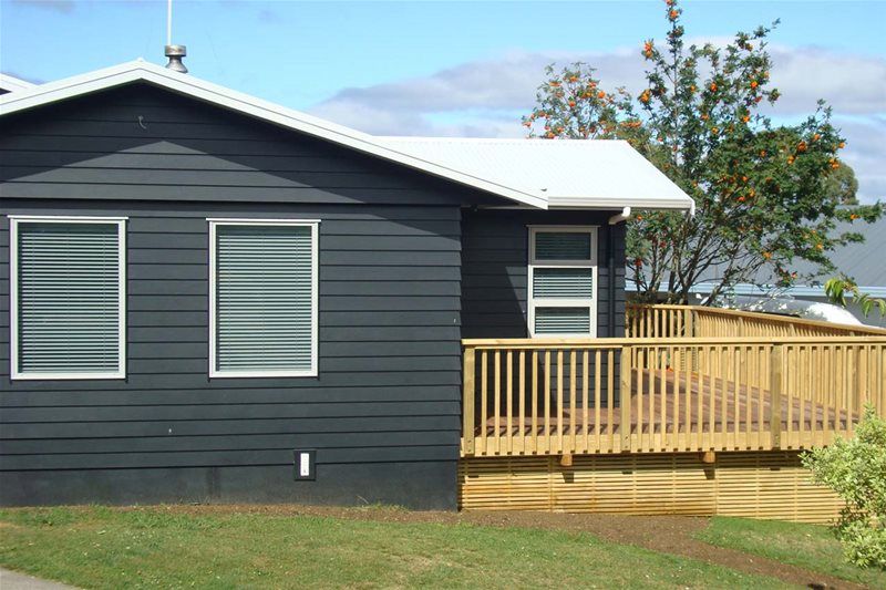 Dark gray house with white-trim windows and a wooden deck on a grassy lot under a blue sky
