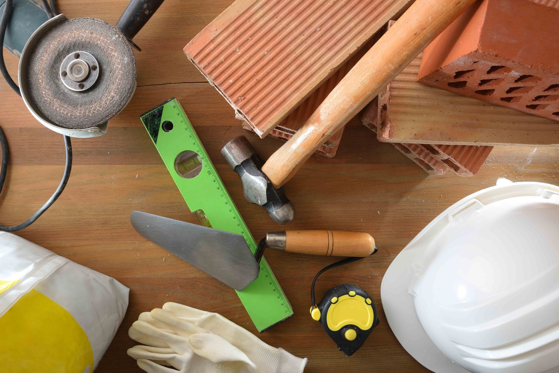Construction tools: grinder, brick, level, trowel, hammer, measuring tape, hard hat, gloves, and cement bag on wooden surface.
