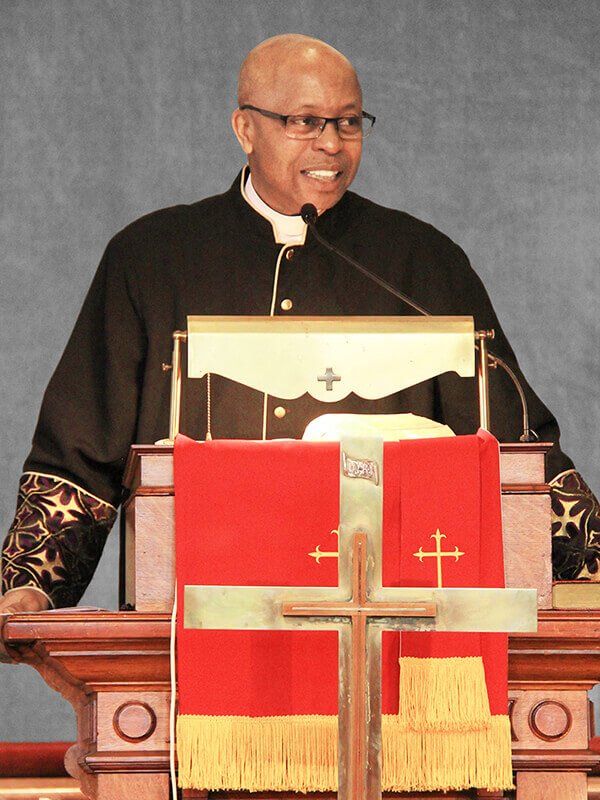 Speaker at a church pulpit with red and gold cloth and a microphone, smiling as he addresses the congregation