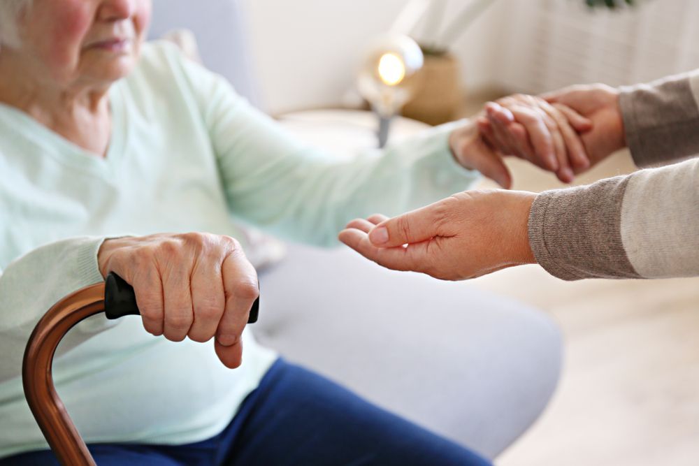 Caregiver offering a hand to an elderly person holding a cane on a sofa