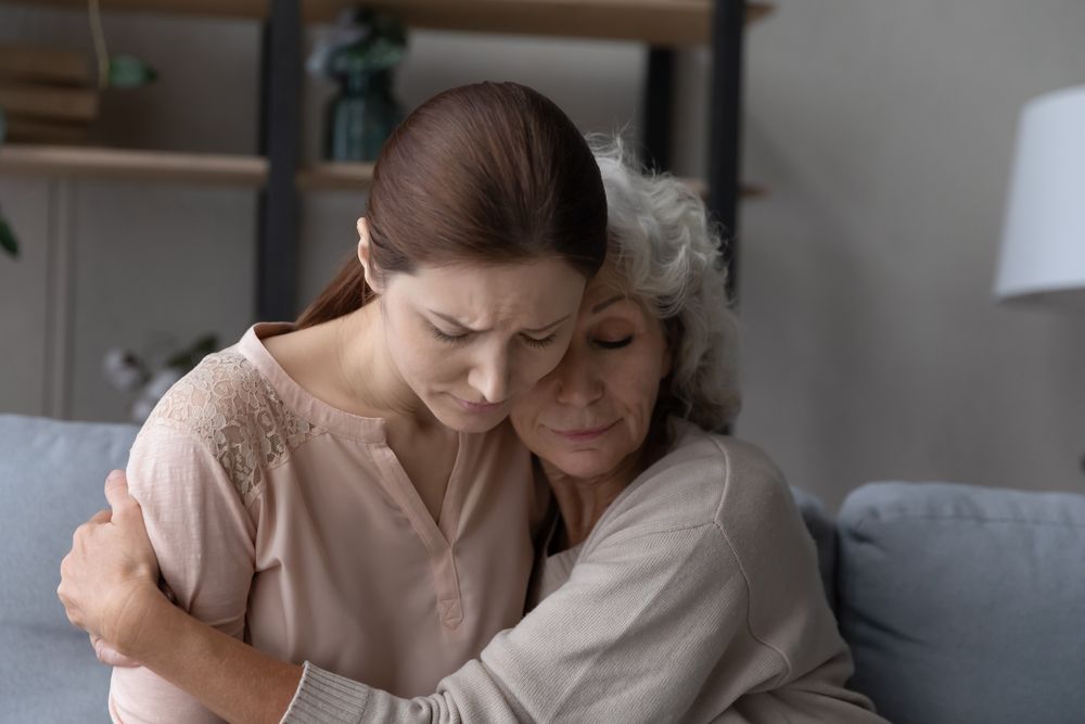 Two women embrace on a couch, one comforting the other in a cozy living room.