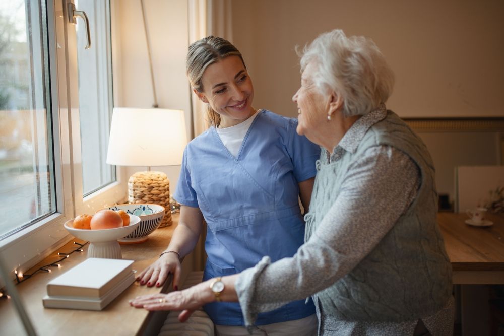 Caregiver in blue scrubs helping an elderly woman stand by a window with books and a lamp nearby