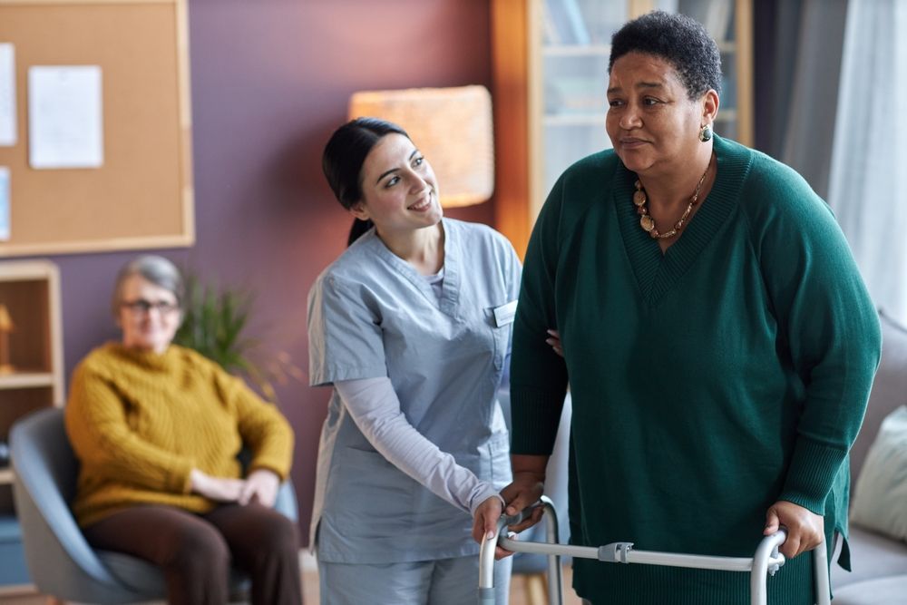 Caregiver assists a patient with a walker in a care facility lounge while another person sits in the background
