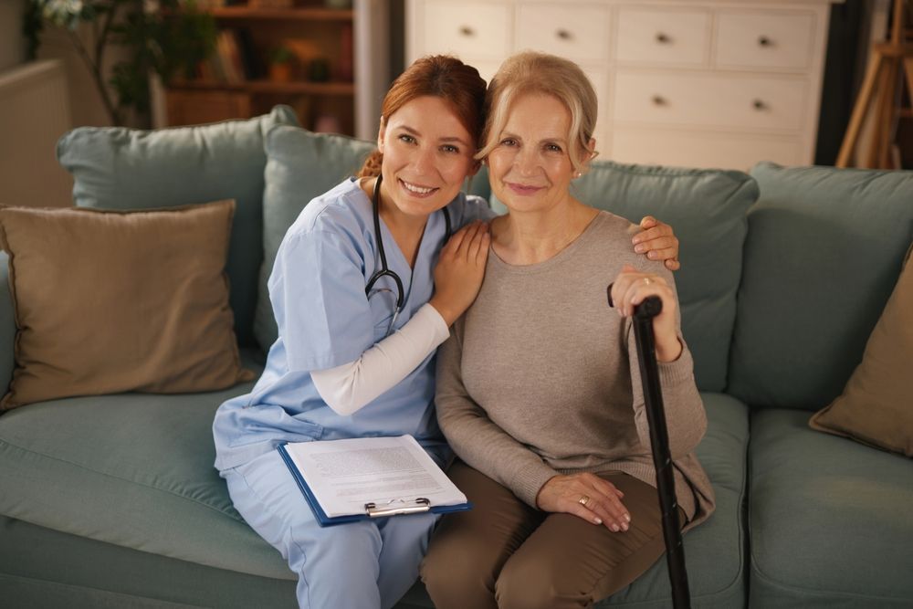 Healthcare worker with clipboard sitting beside a smiling patient on a couch in a living room