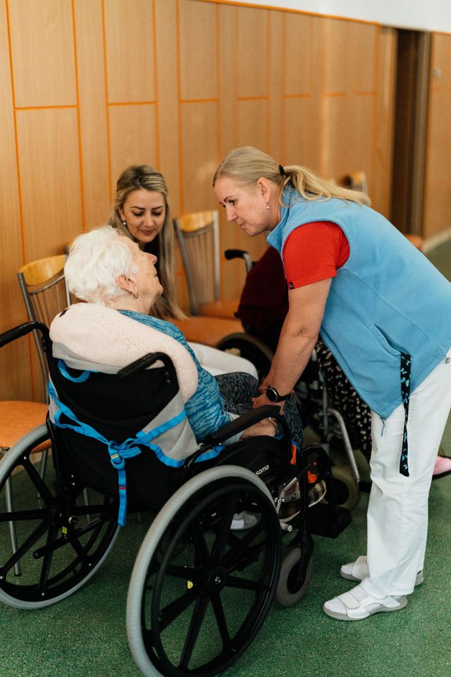 Two women assisting a person in a wheelchair indoors, with one standing beside and one seated nearby.