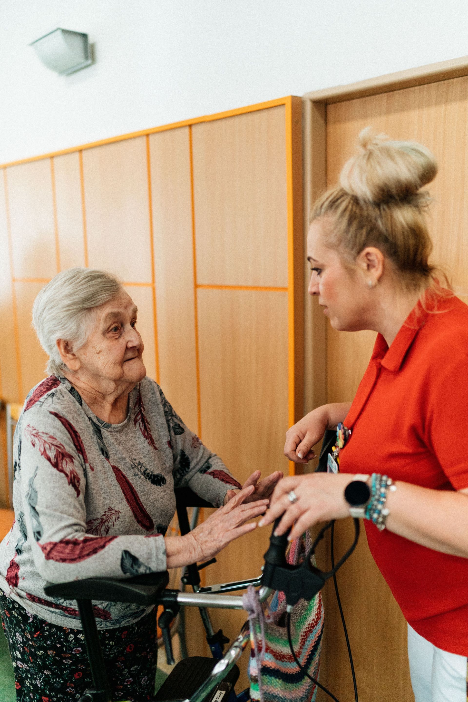 Woman in red shirt assisting elderly woman with a walker in a room with wooden cabinets