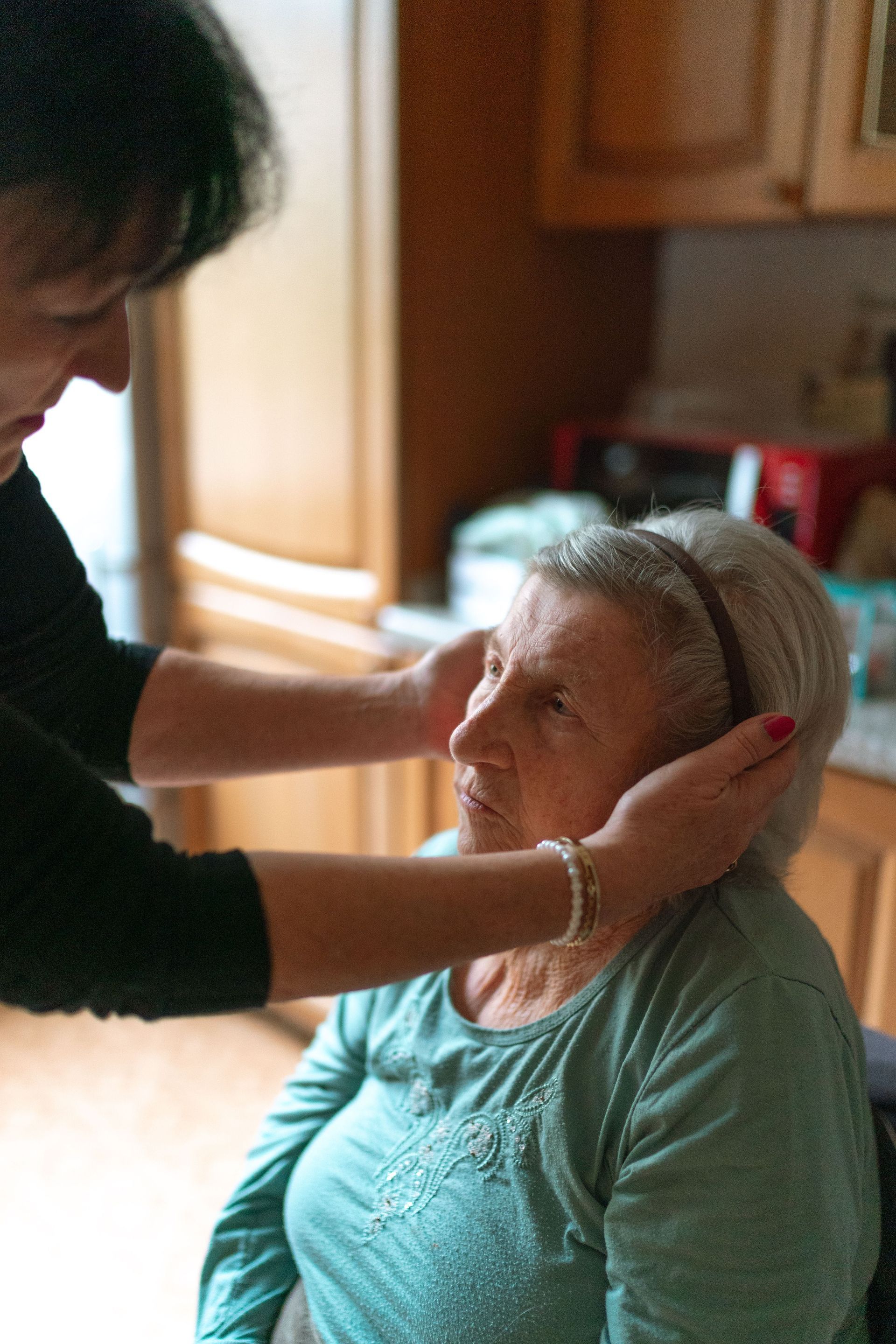Woman adjusting an elderly person's hair in a kitchen, both seated close together.