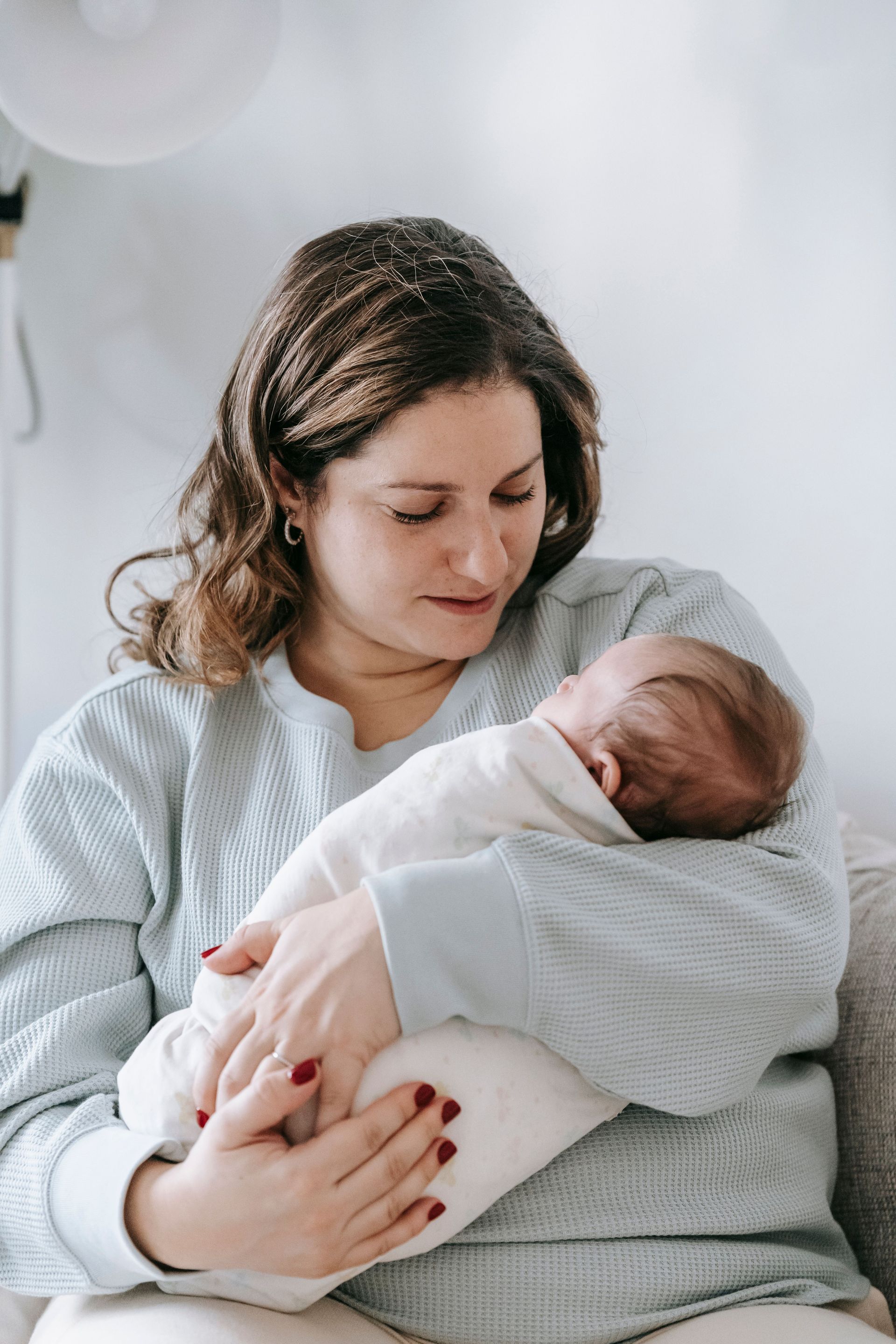 Woman in a gray sweater gently cradling a sleeping baby wrapped in a white blanket
