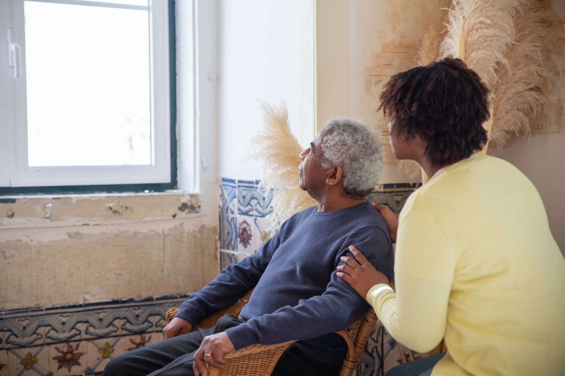 Two people sit by a bright window, looking outside in a cozy room.