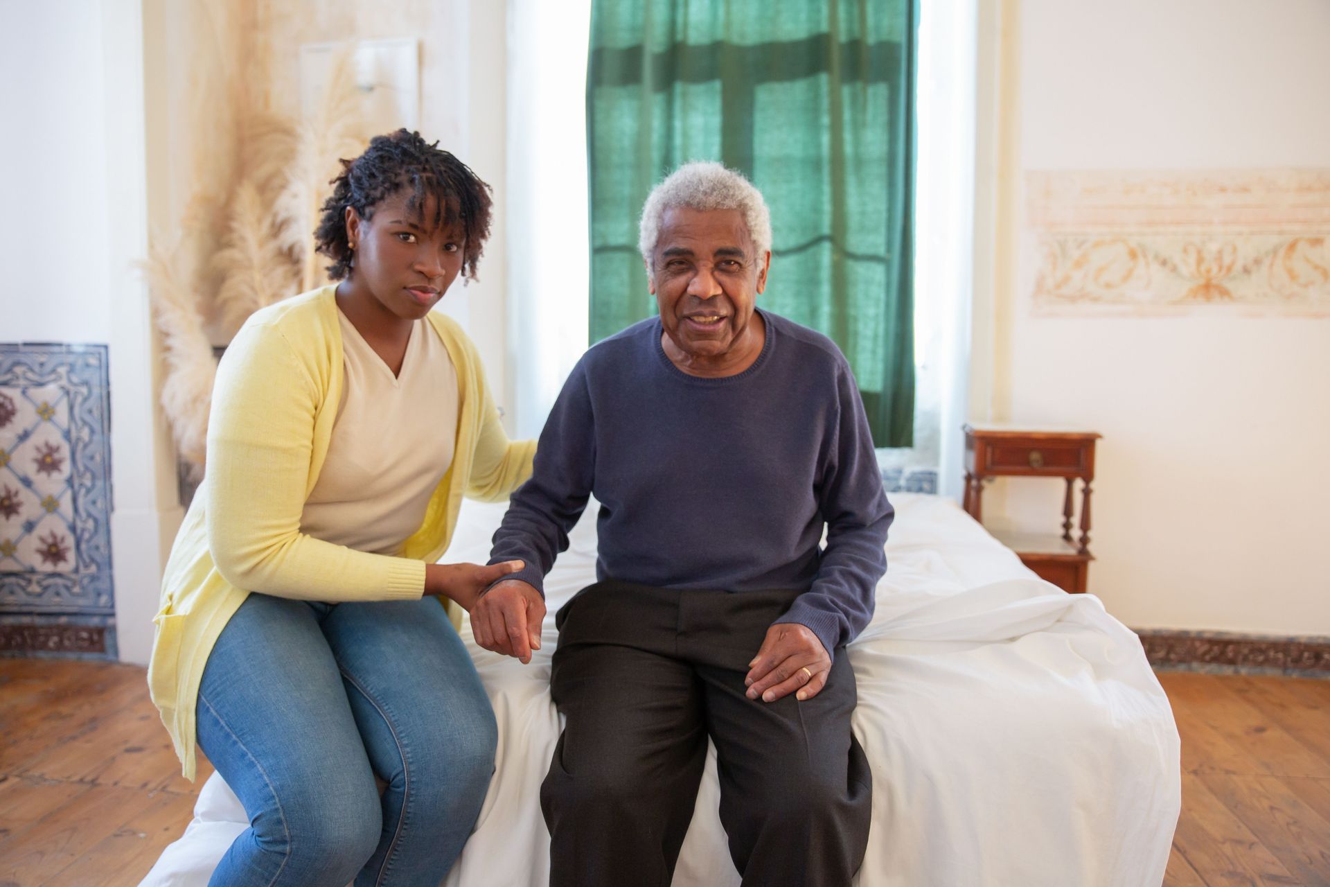 Two people sitting on a bed in a bright bedroom, holding hands and looking at the camera.
