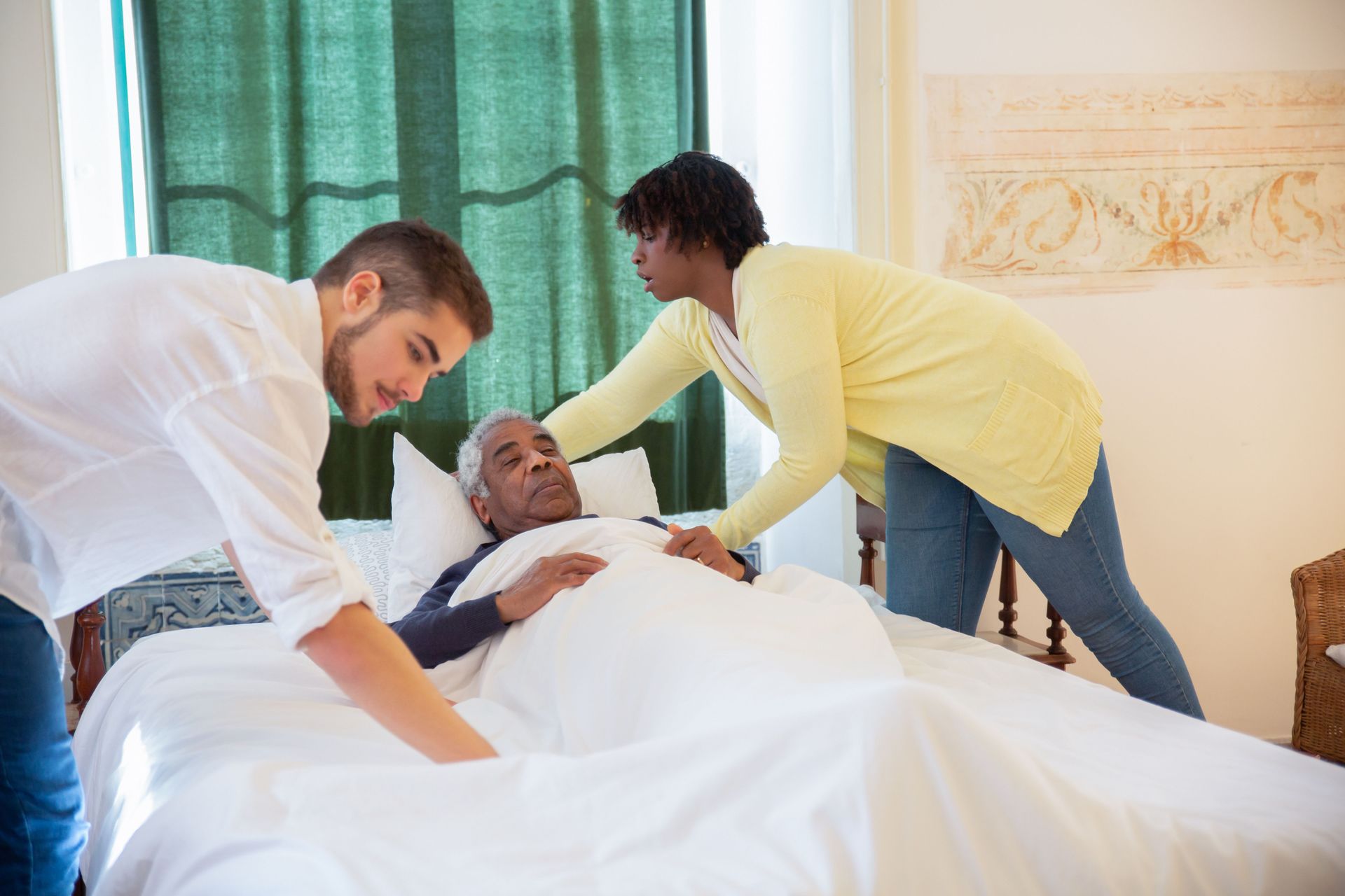 Two caregivers lean over a patient in bed in a bright room with green curtains.