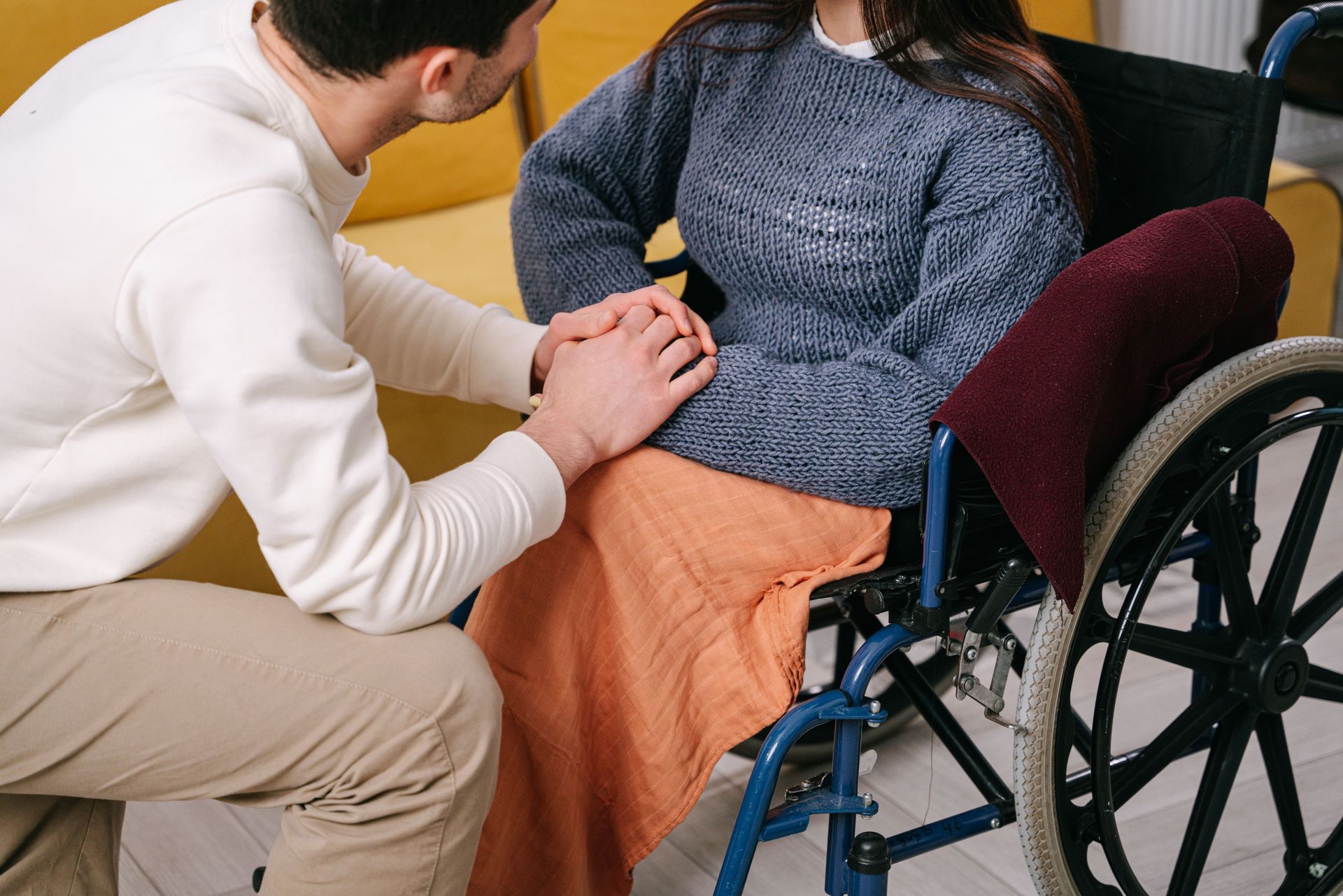 Person kneeling beside a wheelchair user, holding their hands in a caring gesture.