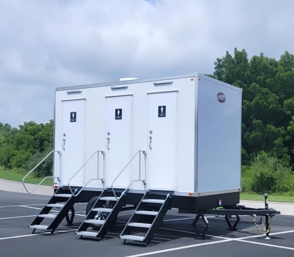 White portable restroom trailer with three stalls, each with stairs and a ramp, parked outdoors.