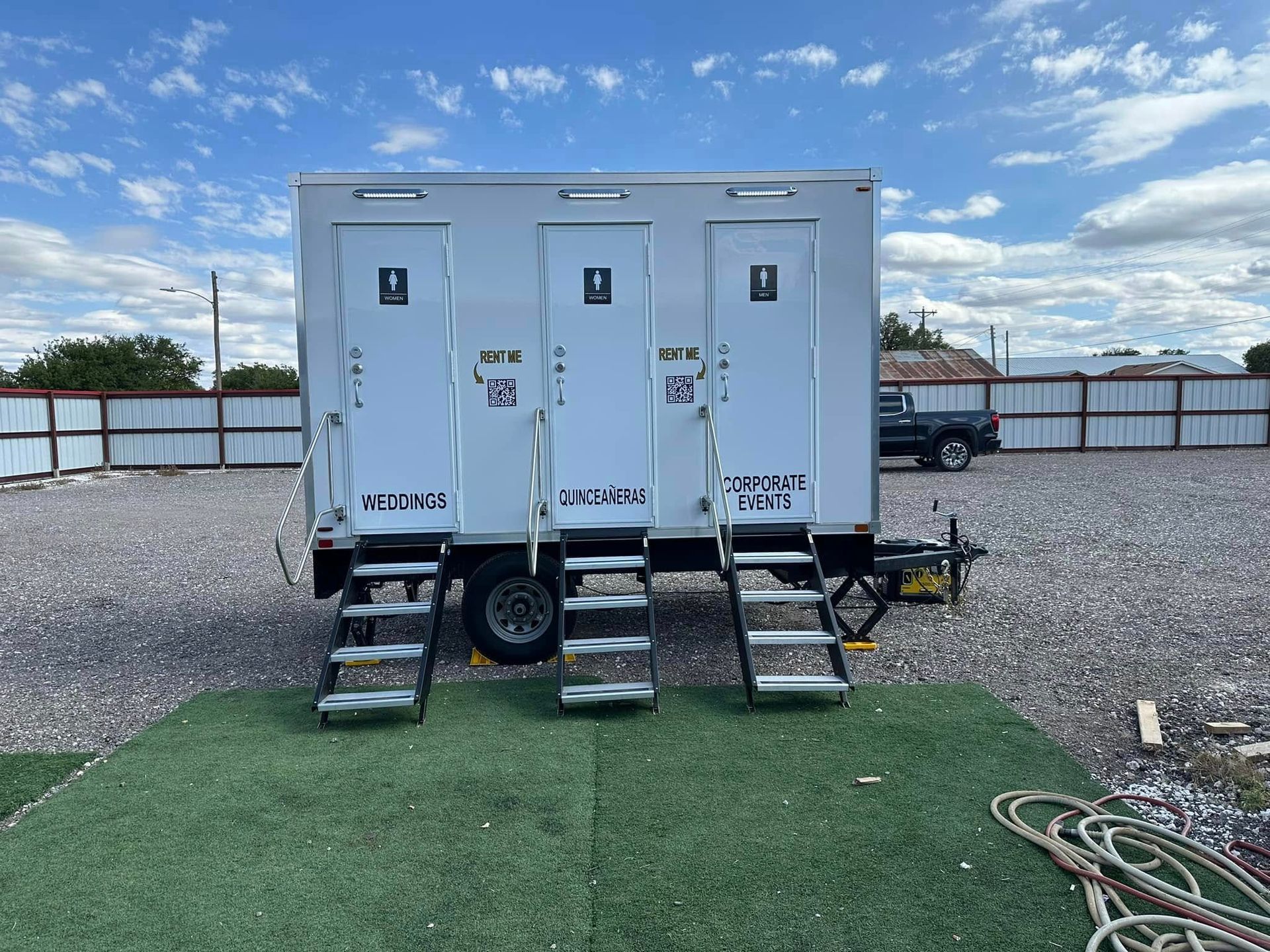 Three-stall portable restroom trailer on a gravel lot. Each door has a handle and step-up stairs.