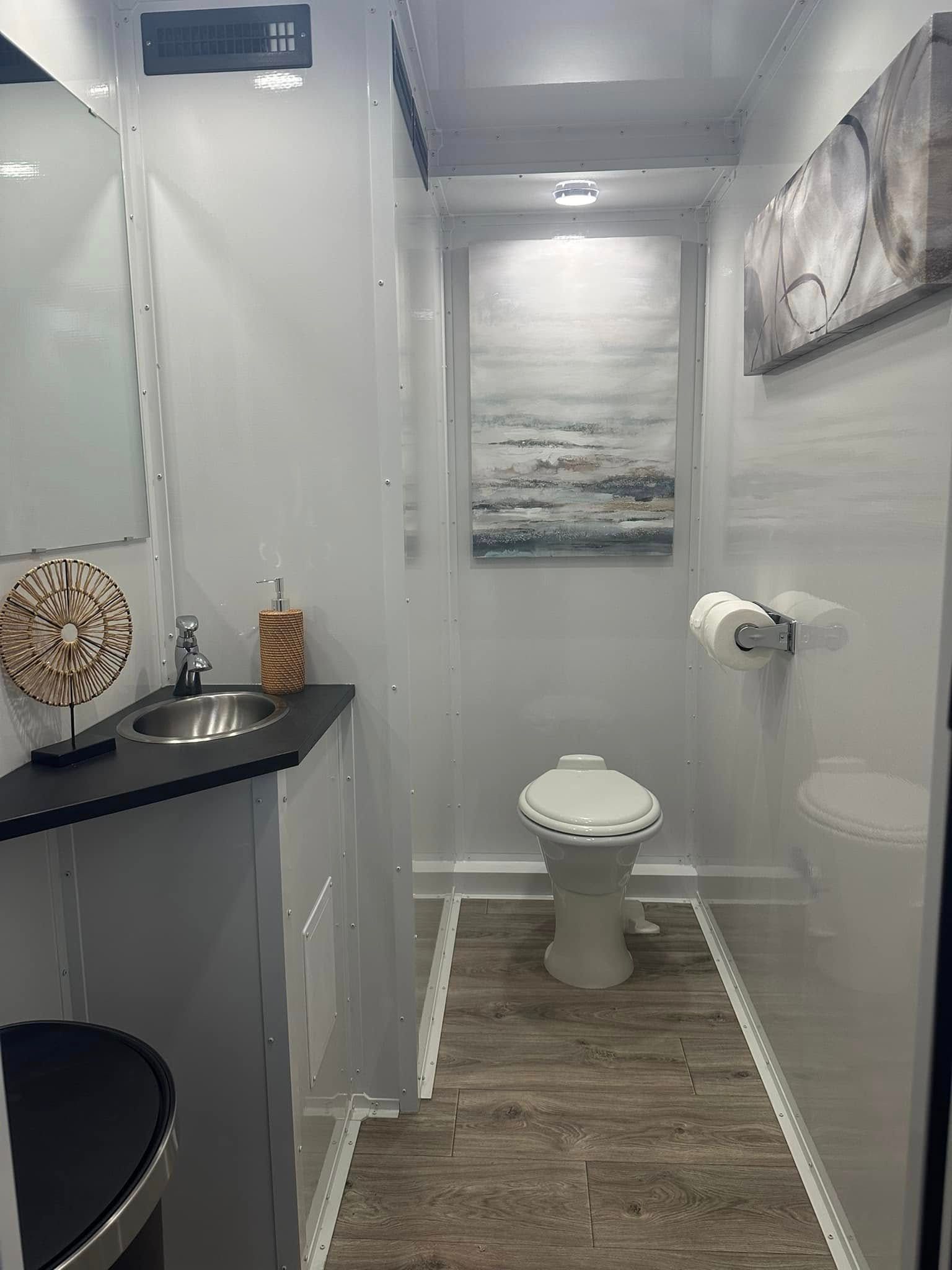 Interior of a luxury portable restroom with a toilet, sink, and artwork. White walls, wood-look floor.