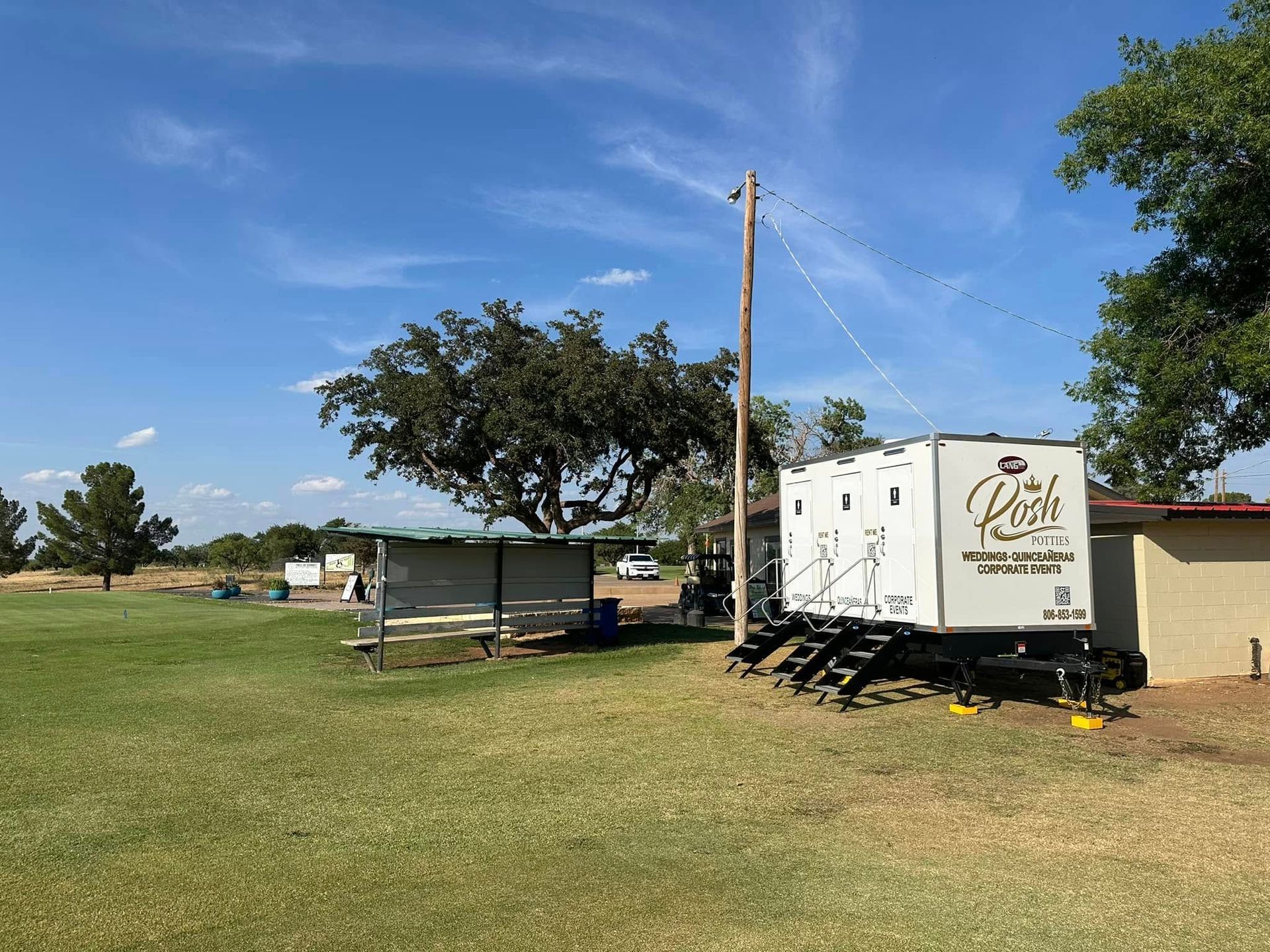 Camping scene with a trailer, shed, and power pole on a grassy field under a blue sky.