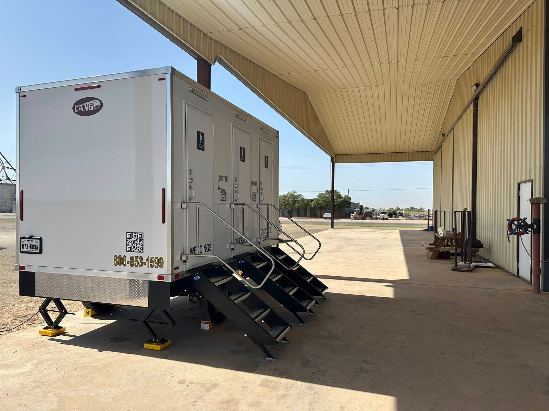 White portable restroom trailer with steps, under a covered area, next to a building.