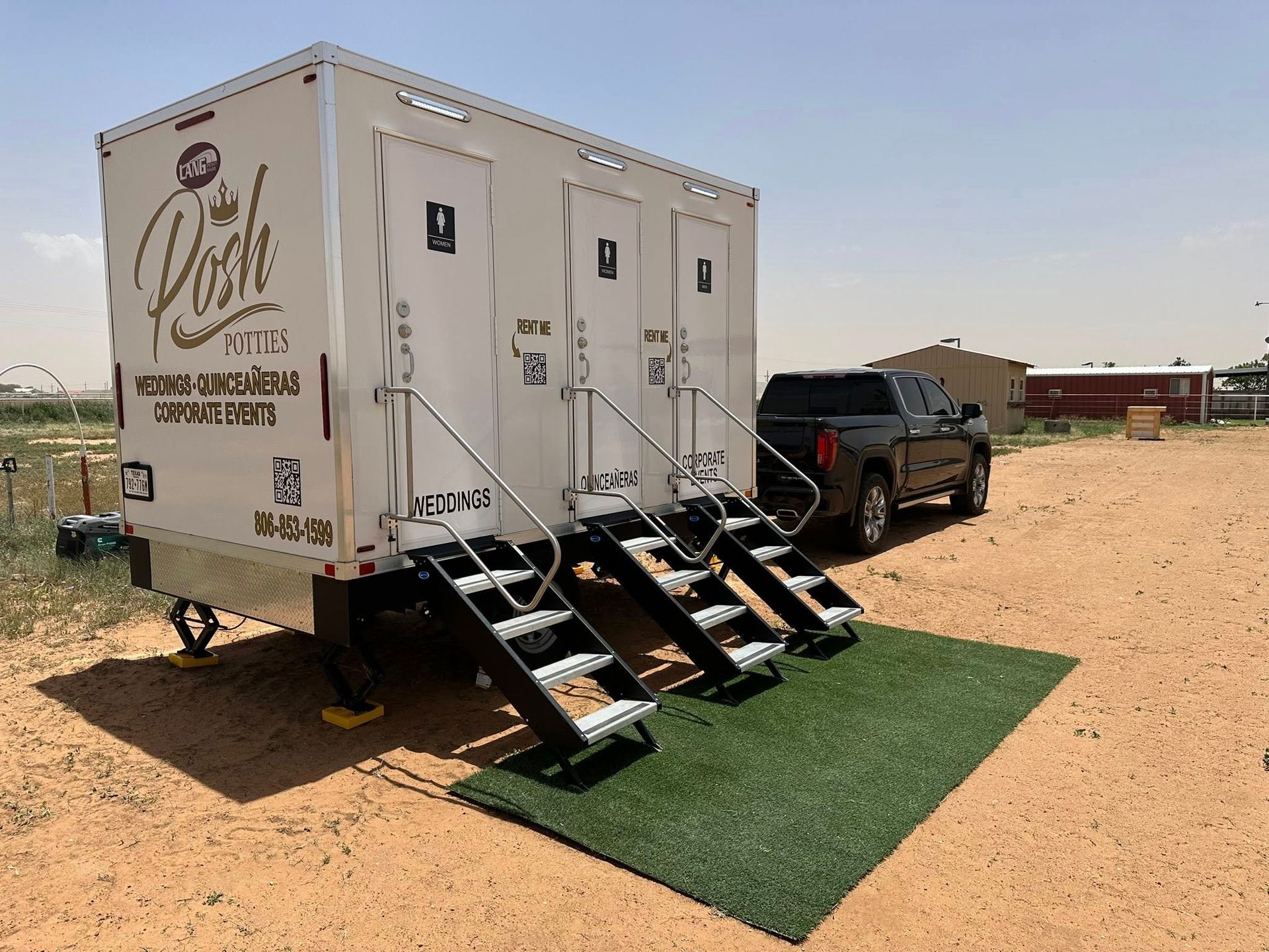 Portable restroom trailer, white with three entrances, steps, parked on dirt with black truck.