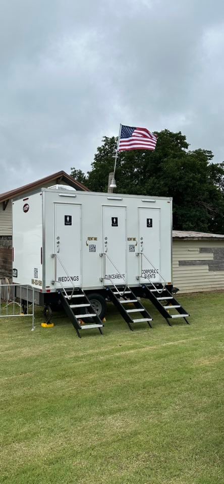 White portable restrooms with stairs, American flag on pole. Outdoors, overcast sky, green grass.