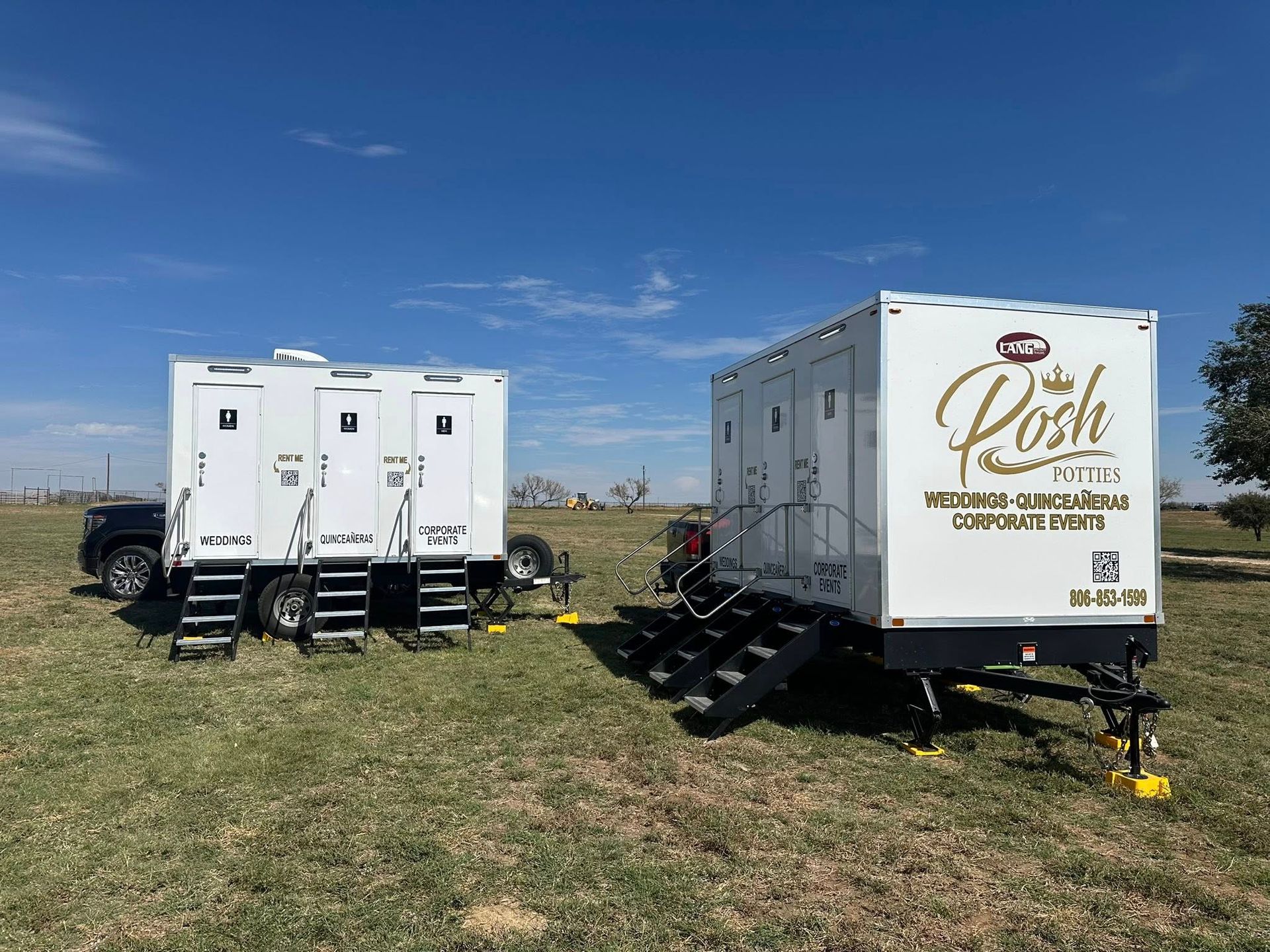 Two white portable restroom trailers on a grassy field under a blue sky. One has 