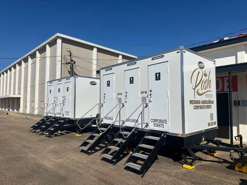 Portable restrooms on trailers. White units with steps and branding. Outdoors, sunny day.