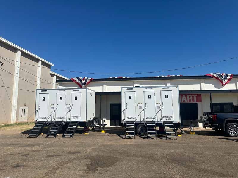 Two white portable restroom trailers parked outside a building under a blue sky, with steps and flags.