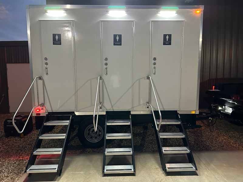 Three-stall portable restroom trailer with steps and handrails. Exterior is white with overhead lights.