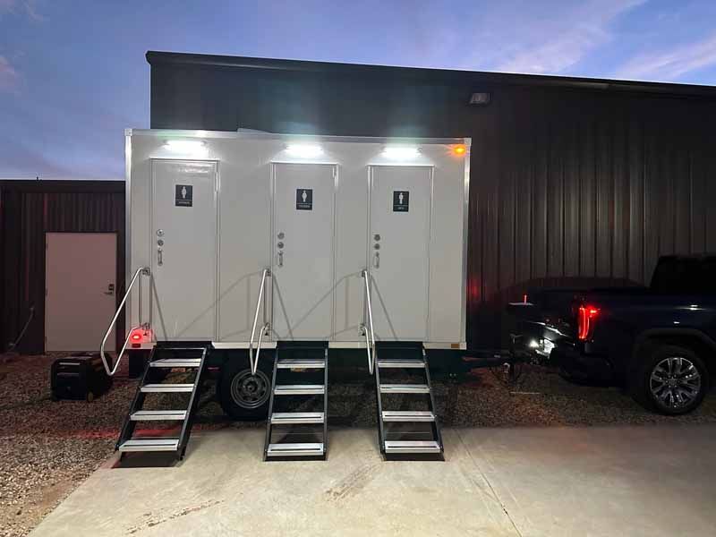 White portable restroom trailer with three individual stalls; black stairs; evening outdoor setting.