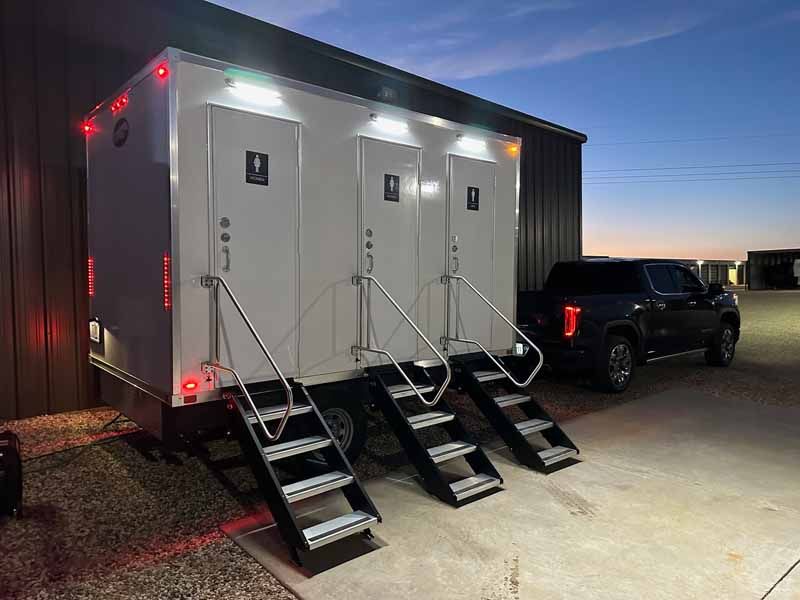 White portable restroom trailer with three doors and steps, parked near a building and a truck.