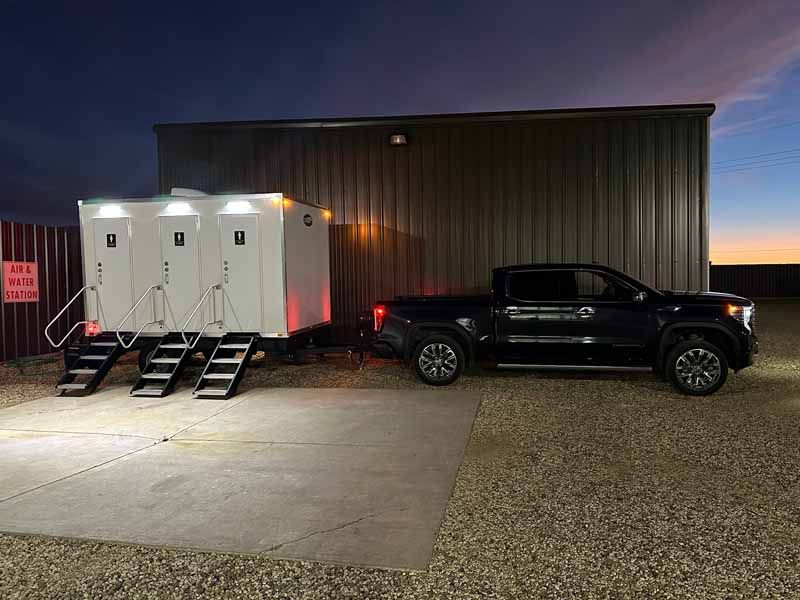 A trailer with three white portable toilets and a black truck parked next to a building at dusk.