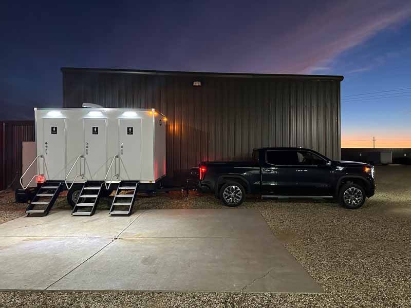 A black pickup truck towing a trailer with four white portable restroom stalls, parked at dusk.