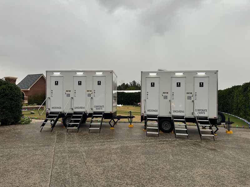 Two white portable restroom trailers on a gray concrete surface, in front of a house, under a cloudy sky.