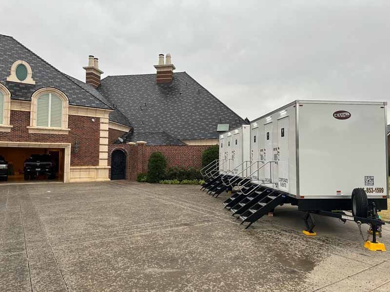 White portable restroom trailer parked on a concrete driveway, next to a brick house with a dark roof.