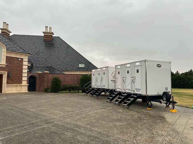 Two white portable restrooms on a trailer parked on a driveway next to a brick building.
