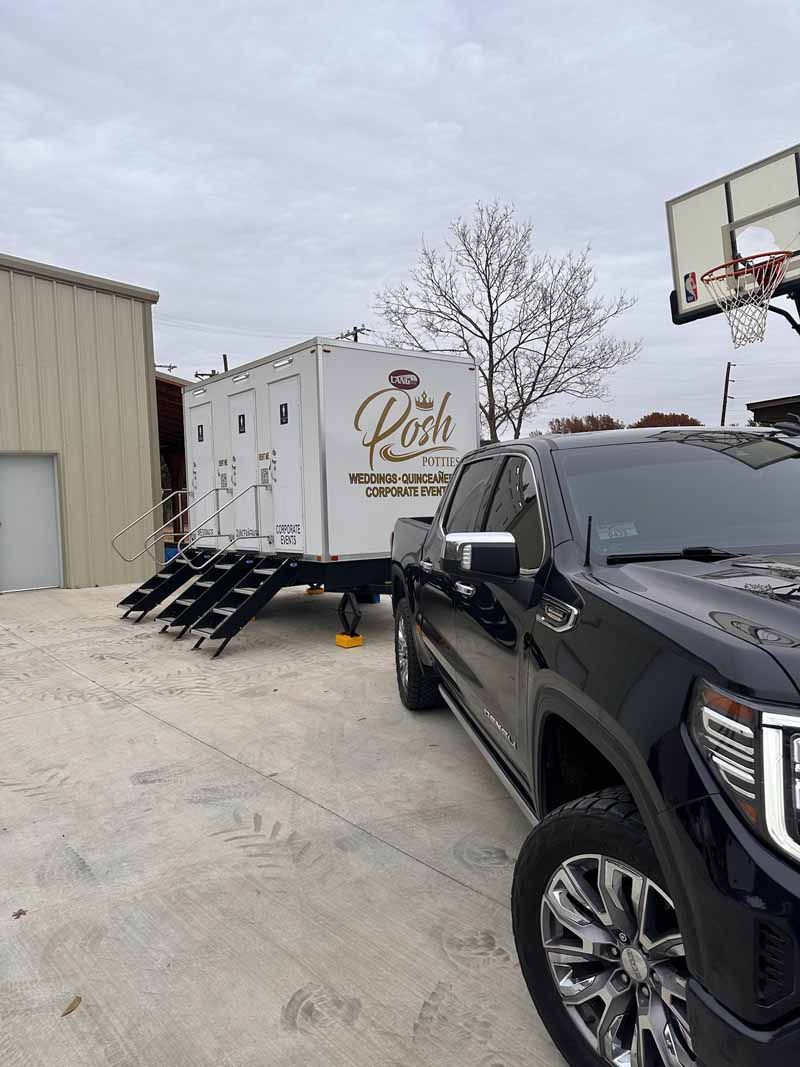 Black truck towing a white portable restroom trailer with gold lettering; parked on concrete.