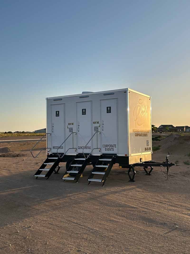 A white portable restroom trailer with three stalls sits on dirt ground with steps.
