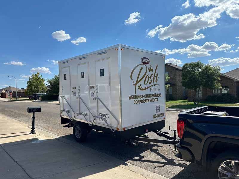 A portable restroom trailer hitched to a truck is parked on a residential street. White exterior with gold lettering.