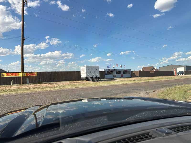 View of a trailer and building behind a wooden fence, on a roadside under a blue sky.