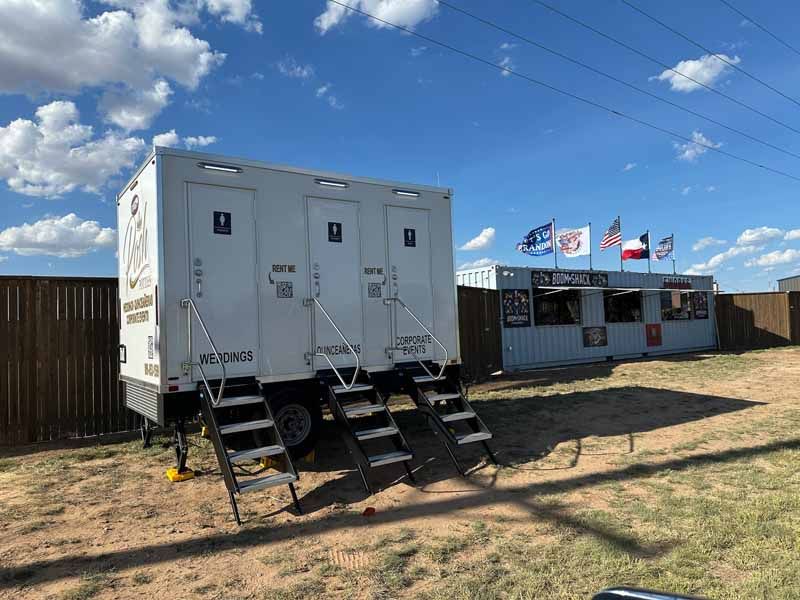 Portable restroom trailer with three stalls and a concession stand with flags on a sunny day.